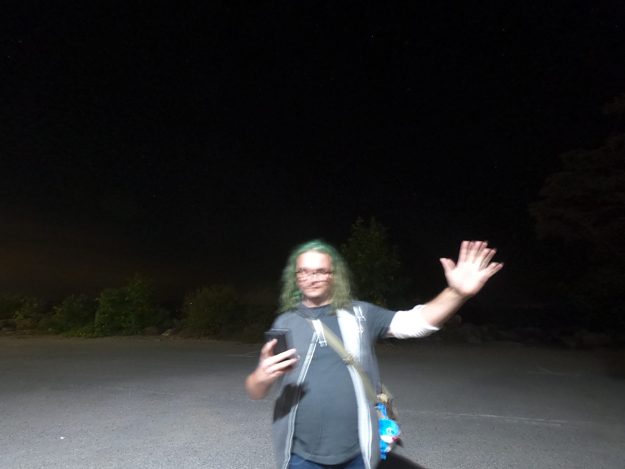 A man with long green hair and no beard stands on a parking lot at night, the photo has motion blur"